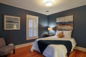 bedroom with furniture, hardwood floors, linavy wall paint, plantation shutters, white comforter, bedside table, bedside lighting, navy throw, art over bed 